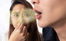 A woman pinches her nose with a distressed expression while a man exhales visibly bad breath. The scene conveys discomfort and awkwardness