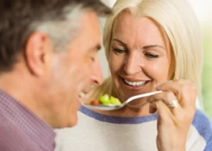 A smiling woman holds a spoon of vegetables offering it to a man