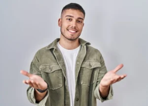 A smiling young man gesturing with open hands against a plain background.