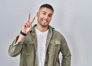 Young man smiling showing a peace sign with his right hand. Neutral background, relaxed and friendly mood.