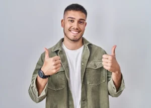 A smiling man in a green jacket and white shirt gives a thumbs-up with both hands