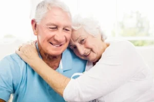 An elderly couple embraces warmly on a sofa. The man in a blue shirt smiles, while the woman in white rests her head on his shoulder, conveying love and comfort.