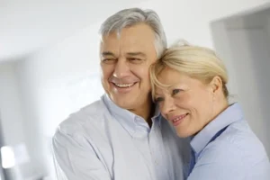 A smiling older couple embraces, conveying warmth and happiness. They wear light-colored shirts, standing in a softly lit, neutral-toned room.