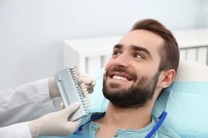 A smiling man in a dental chair gets his teeth color-matched with a shade guide by a dentist. The tone is professional and reassuring.