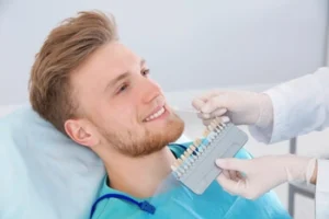 A man with a beard in a dental chair is smiling as a dentist matches tooth shades. Gloves, dental tools, and a calm, clinical setting are visible.
