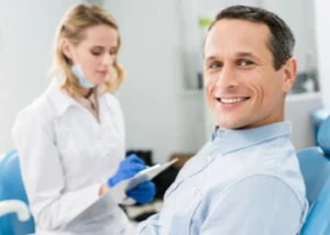 Smiling man sits in a dental chair, wearing a light shirt. A female dentist stands beside him, holding a clipboard and wearing gloves and a mask.