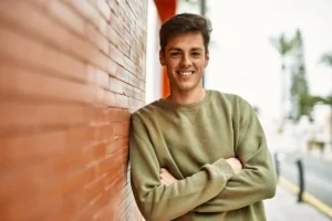 A young man leaning against a red-brick wall with his arms crossed, confidently showcasing his finished smile after a composite veneers procedure