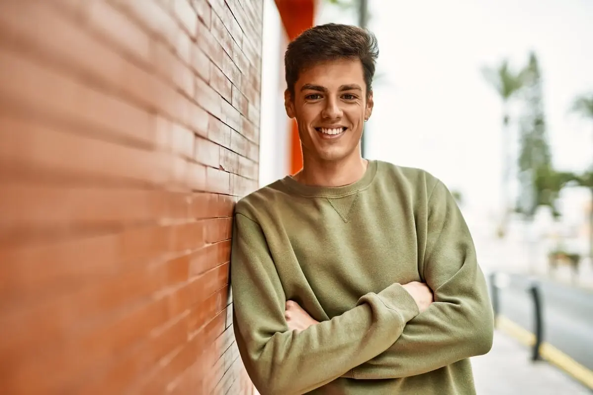 A young man leaning against a red-brick wall with his arms crossed, confidently showcasing his finished smile after a composite veneers procedure