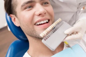 Man sitting in a dental chair, smiling, while a dentist holds a shade guide to his teeth for color matching. The atmosphere is professional and calming.