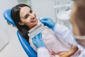 A smiling woman sits in a dental chair as a dentist in gloves matches her teeth shade with a color palette, conveying a friendly, professional atmosphere