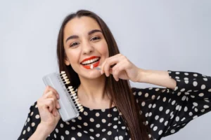 A smiling patient holding a dental shade guide to her teeth, demonstrating the preparation stage for veneers where the perfect tooth color is selected for a custom, natural-looking finish.