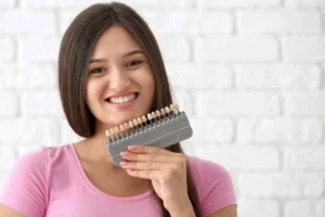 A smiling patient holding a dental shade guide to her teeth, highlighting the aesthetic pros and cons of choosing the right color and translucency for porcelain veneers.