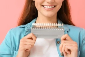 Smiling woman holding a dental shade guide against a pink background. She is wearing a blue shirt, conveying a sense of positivity and dental care