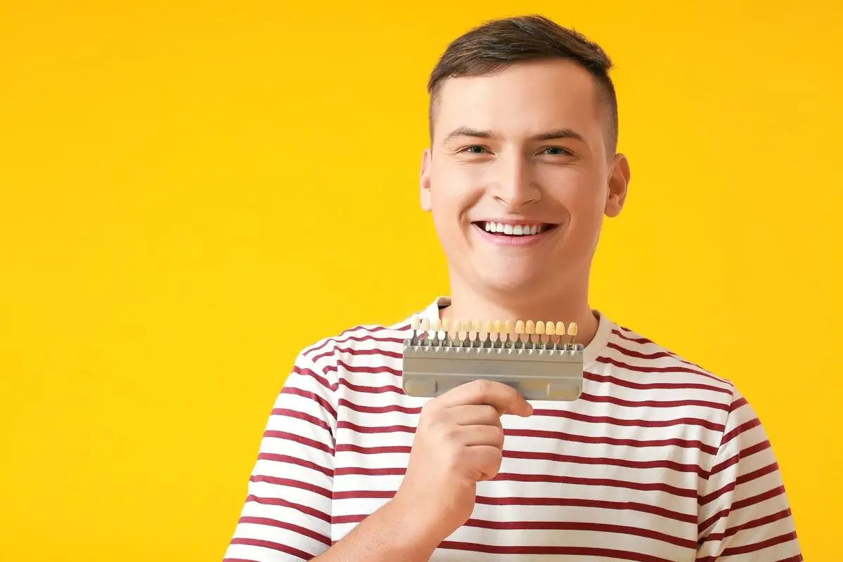 Smiling person holding a dental shade guide against a vibrant yellow background, wearing a white and red striped shirt, conveying a cheerful mood.