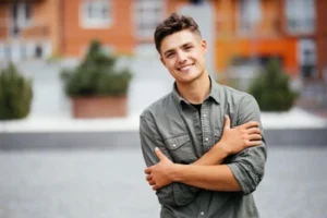 Smiling young man in a green shirt stands with arms crossed in a courtyard, with a blurred background of brick buildings and greenery, conveying warmth and approachability.