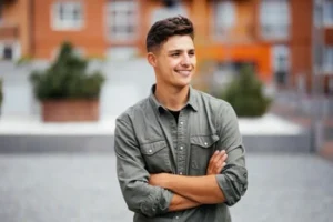 Young man in a green shirt stands outdoors with arms crossed, smiling confidently. Background features blurred urban setting with warm-toned buildings.