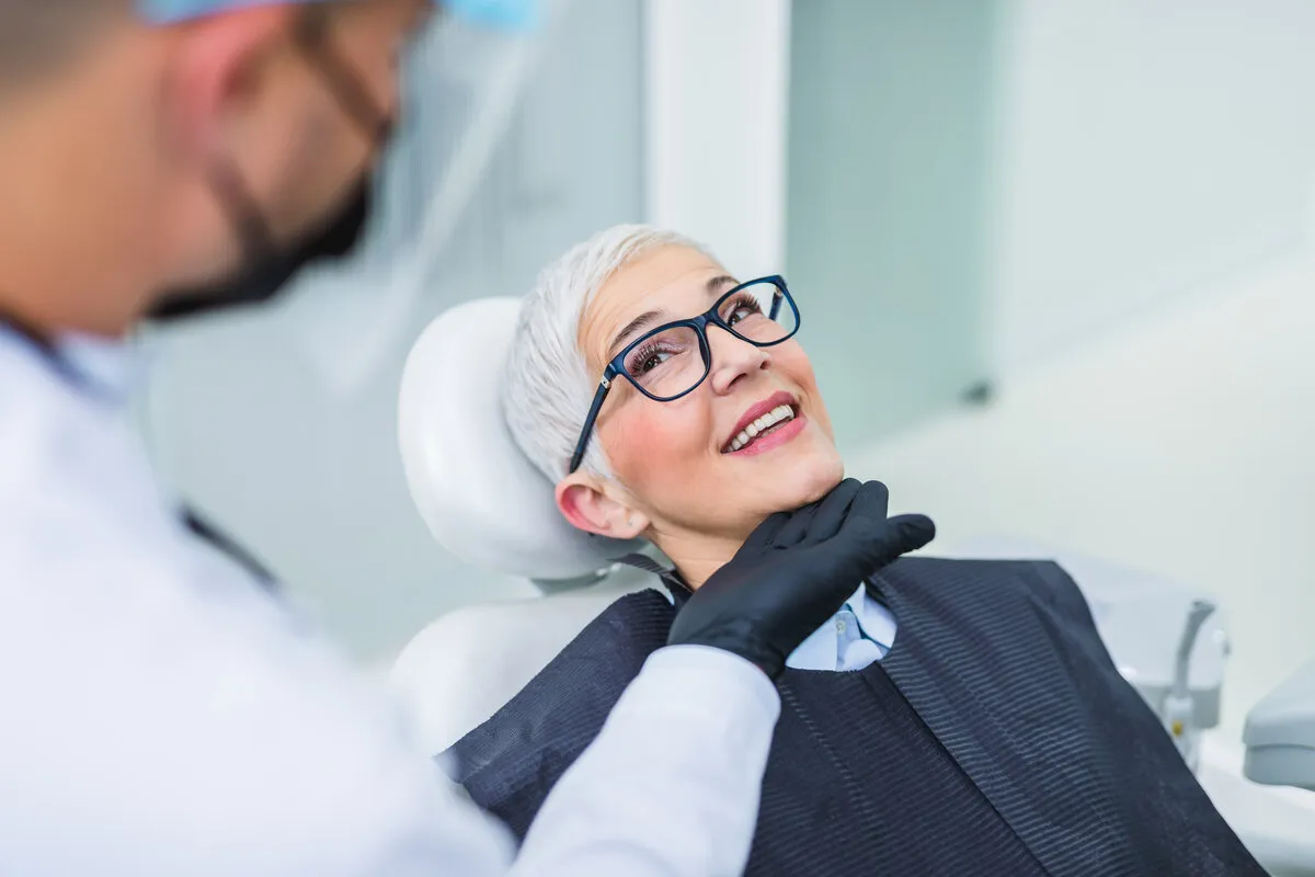 An older woman with short white hair and glasses smiles in a dentist's chair, while a dentist in gloves gently examines her, conveying comfort.
