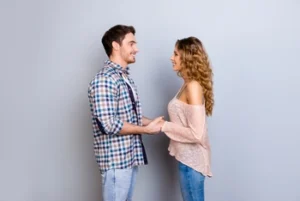 A man and woman stand facing each other, holding hands and smiling warmly.