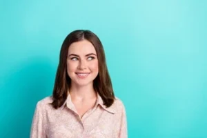 A woman smiles slightly and looks to the side against a bright turquoise background. She's wearing a light pink collared shirt.