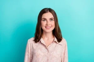 Young woman wearing a light pink shirt, smiles brightly against a vibrant turquoise background. The mood is cheerful and welcoming.