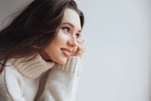 Woman in a cozy sweater smiles thoughtfully, resting her chin on her hand. She gazes to the right, exuding warmth and contemplation against a soft, light background.