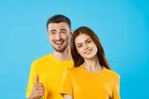 A smiling man and woman in yellow shirts against a blue background, with the man giving a thumbs-up to represent patient satisfaction and the success of professional dental treatment.