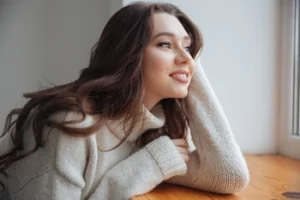 Young woman with long brown hair wearing a cozy sweater, gazing out a window.