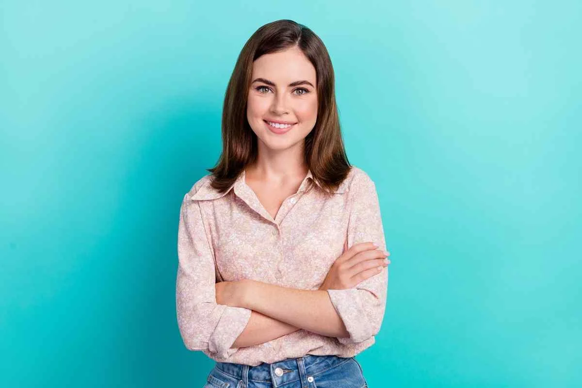 Smiling woman stands with arms crossed against a bright turquoise background. She exudes confidence and warmth.