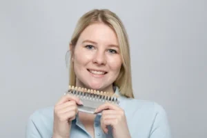 A smiling person with blonde hair holds a dental shade guide against a light gray background, conveying a sense of warmth and professionalism.
