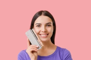 A smiling woman holds a dental shade guide next to her face against a pink background. She wears a purple shirt, conveying a cheerful and bright mood