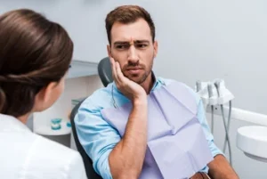 A man sitting in a dental office looks concerned, holding his cheek with one hand. He wears a dental bib, and a dentist is seated opposite him.