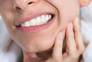 Close-up of a person showing teeth while pressing a hand against the cheek, indicating possible dental pain or discomfort. The expression suggests unease.