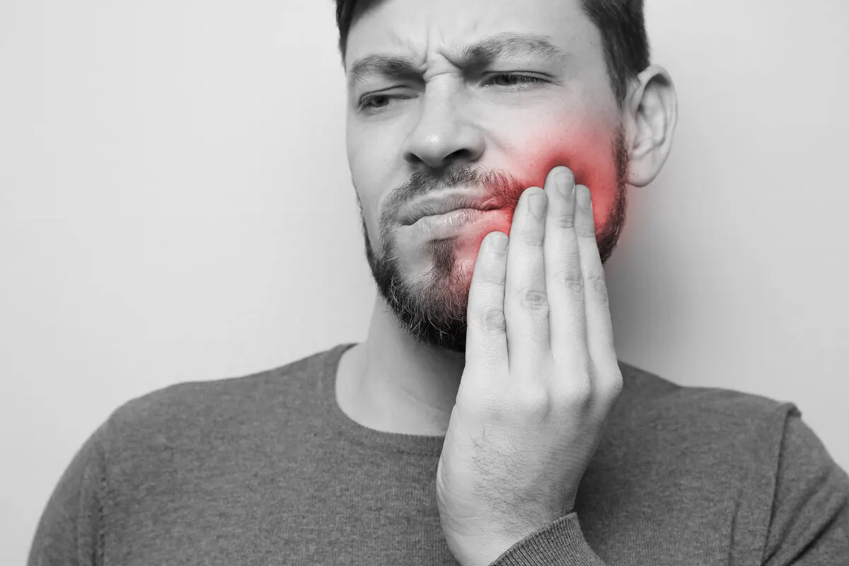A man with a beard grimacing and holding his jaw, with a red highlighted area indicating sharp tooth pain or a dental emergency that requires immediate professional attention