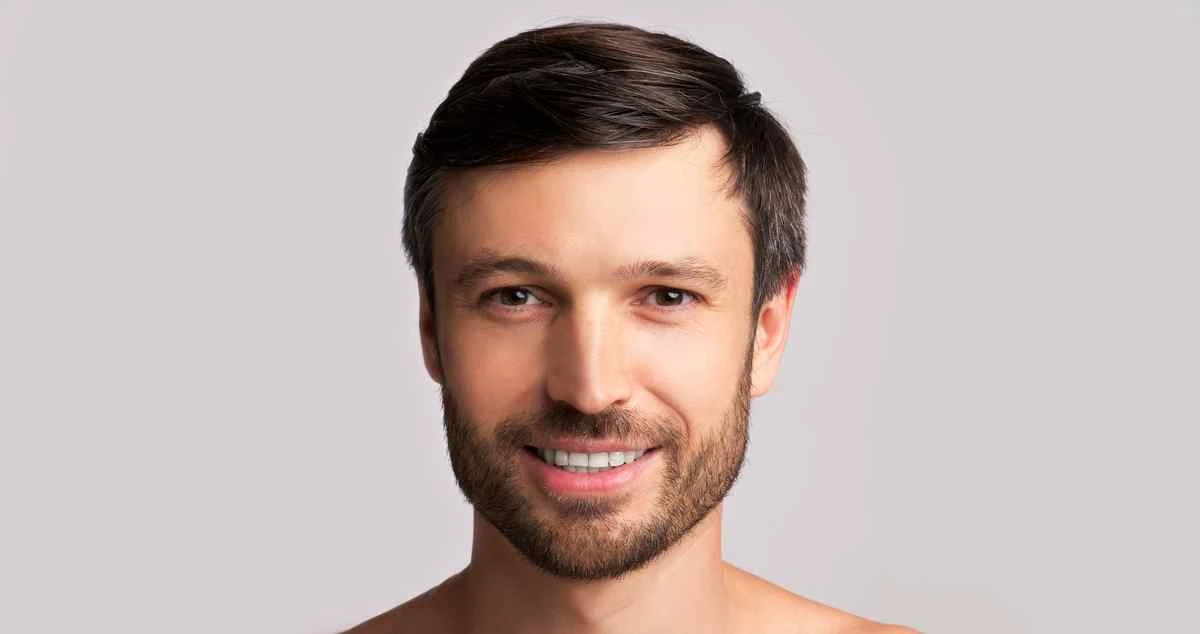 A smiling man with a neat beard and short dark hair, showcasing a healthy and confident smile that reflects the success of routine dental care