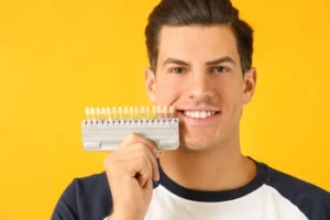 Smiling man holding a dental shade guide near his mouth against a bright yellow background, showcasing teeth color comparison for dental treatment.