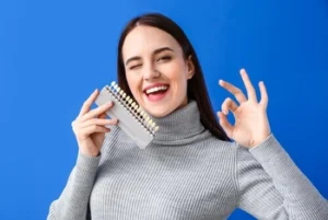 Smiling person winks, holding a teeth color chart and giving an OK sign against a bright blue background