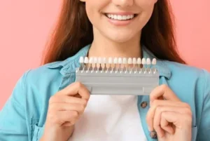 A smiling woman holds a dental shade guide against a pink background. She wears a blue shirt, showcasing her bright, aligned teeth