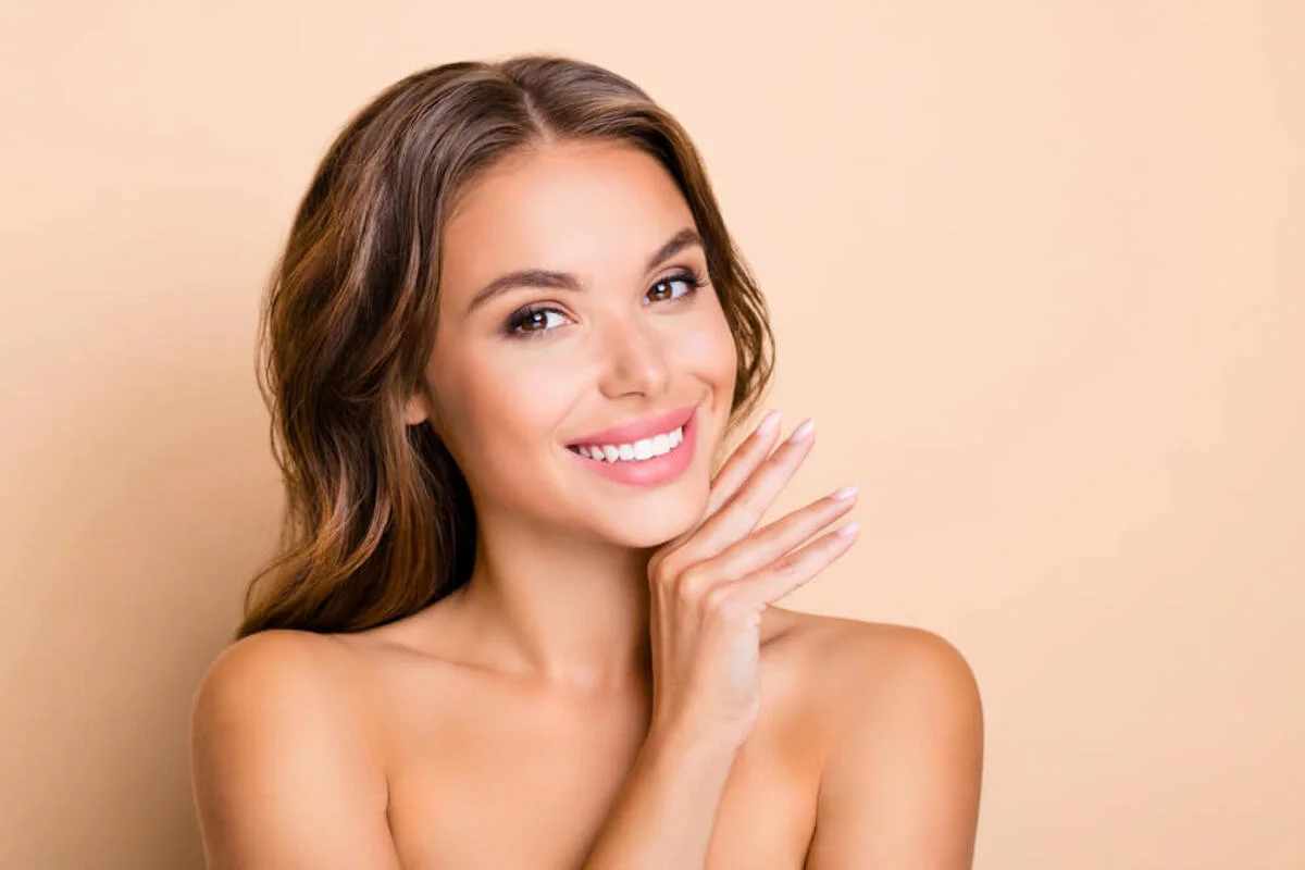 A smiling woman with a radiant complexion posing against a soft beige background, highlighting the aesthetic beauty and self-confidence achieved through professional cosmetic dentistry