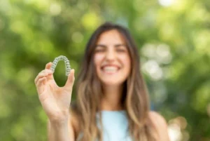 A smiling woman holding a clear dental aligner, showcasing the discreet and lifestyle-friendly nature of orthodontic treatment with a natural, blurred outdoor background