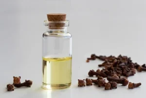 Small glass bottle with cork lid containing pale yellow oil, placed beside a scattered pile of dried cloves on a white surface