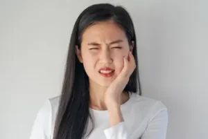 A woman with long dark hair holds her cheek and winces in pain. Her expression conveys discomfort, possibly a toothache.