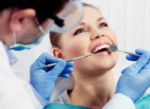 A woman in a dental chair smiles as a dentist in gloves examines her teeth with a mirror tool. Bright, clinical setting conveys a sense of care and professionalism.