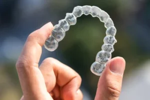 A hand holds a clear dental aligner against a blurred background. The transparent device is curved, showcasing its detailed structure and smooth surface.
