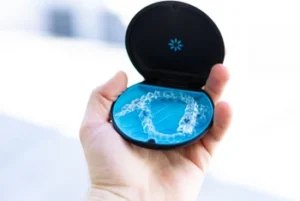 A hand holds a black case with a blue interior containing a clear dental retainer. The image conveys a sense of cleanliness and dental care focus.