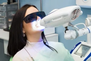 A woman wearing protective glasses undergoes teeth whitening at a dental office. A bright light is directed at her mouth