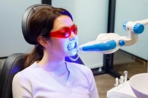 Woman in dental chair wearing red protective glasses undergoing teeth whitening with a blue LED light device