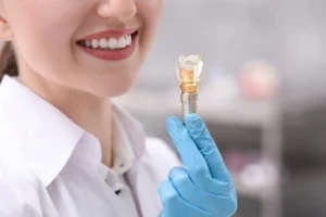 A person in a white coat smiles while holding a dental implant with a gloved hand
