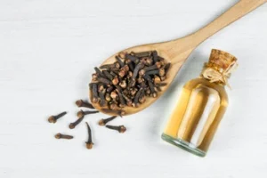 A wooden spoon filled with dried cloves is placed on a light surface, next to a small corked glass bottle of clove oil, conveying a natural, aromatic scene