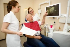 An elderly man in a dental chair points at an X-ray on a monitor. Two dental professionals beside him discuss, holding a chart.