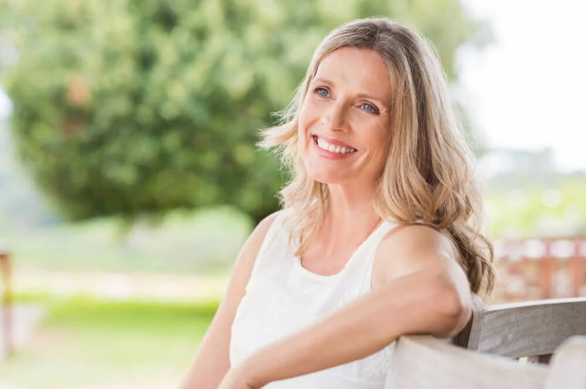 Smiling woman with long blonde hair sits on a park bench, wearing a white sleeveless top